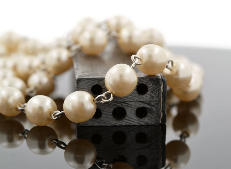 May include: A string of faux pearls draped over a black rectangular object with holes. The pearls are cream coloured and the object has a rough texture.