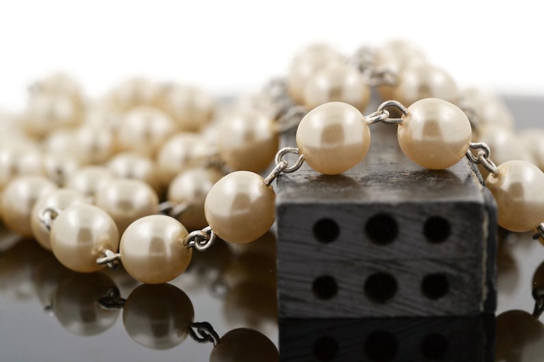May include: A strand of white faux pearl beads with silver metal connectors, resting on a black rectangular block with six holes.