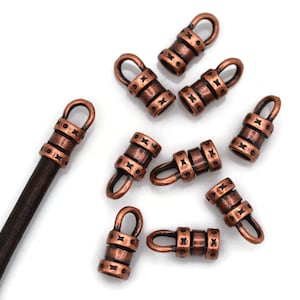 May include: A collection of copper-toned metal cord ends featuring a cylindrical shape with a loop at the top and decorative etched star patterns. One cord end is attached to a dark brown leather cord, while the others are scattered around.