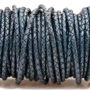 May include: A spool of braided leather cord in a deep teal color. The cord is tightly woven, creating a textured surface. The spool is black, and the cord is wound neatly around it, ready for use in jewelry making or crafts.