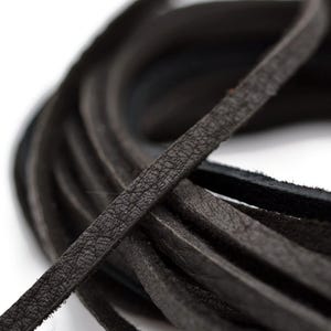 May include: Close-up of a coil of dark gray leather strips. The leather has a textured surface and appears to be flat and narrow. The strips are arranged in a circular pattern against a white background.