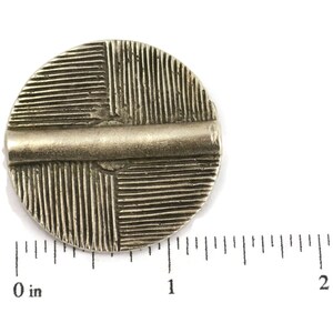 May include: A circular silver-toned metal button featuring a raised horizontal bar across the center and a textured pattern of vertical and horizontal lines in the four quadrants. A ruler shows the button is approximately 1.25 inches in diameter.