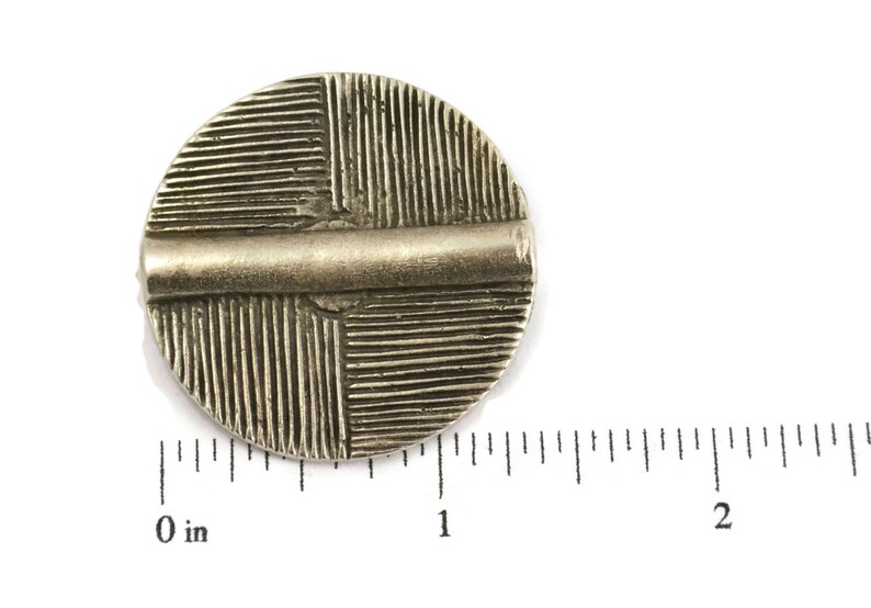 May include: A circular silver-toned metal button featuring a raised horizontal bar across the center and a textured pattern of vertical and horizontal lines in the four quadrants. A ruler shows the button is approximately 1.25 inches in diameter.