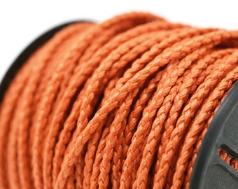 Puede incluir: Un carrete de cordón trenzado de color naranja vibrante enrollado firmemente alrededor de una bobina de plástico negro. El cordón presenta una textura tejida intrincada y consistente en toda su longitud, con un color brillante y flexible.