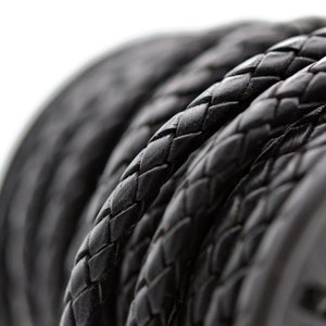 May include: Close-up of a spool of black braided leather cord. The cord is tightly woven, creating a textured surface. The image is set against a white background, highlighting the dark color and intricate pattern of the leather.