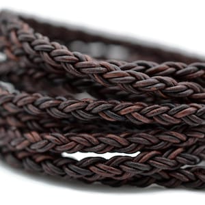 May include: A dark brown braided leather cord coiled in multiple loops. The cord features a tight, round, woven texture with visible variations in the brown tones throughout the material, creating a consistent pattern along the entire length.