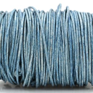 May include: A spool of round leather cord in a mottled light blue and white finish. The cord is wrapped tightly around a cylindrical spool, displaying a textured, weathered appearance with varying shades of blue throughout the length of the material.