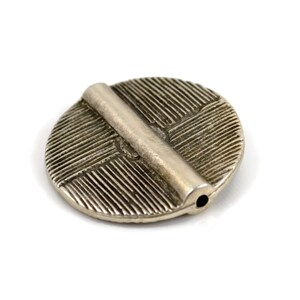 May include: A circular silver-toned metal bead featuring a raised horizontal bar across the center. The surface of the disc is textured with fine, parallel etched lines radiating in different directions. A small hole is visible at the end of the bar.