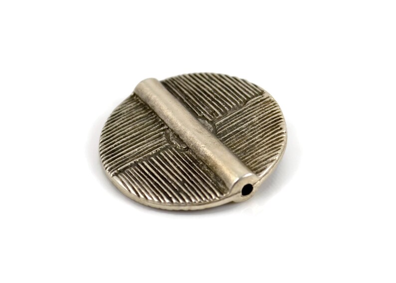 May include: A circular silver-toned metal bead featuring a raised horizontal bar across the center. The surface of the disc is textured with fine, parallel etched lines radiating in different directions. A small hole is visible at the end of the bar.