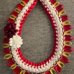 May include: A handcrafted lei necklace featuring a braided design of red, white, and yellow ribbons. The lei is adorned with a burgundy and white flower accent, and a burgundy bow at the top. This traditional Hawaiian garland is a symbol of love and celebration.