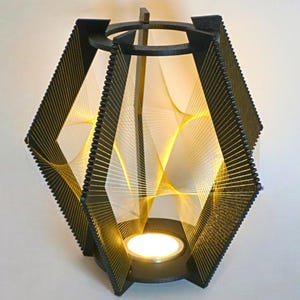 May include: A black geometric lantern with a candle inside. The lantern has a diamond shape with a black frame and gold string design. The candle emits a warm, yellow glow, creating a cozy ambiance. The lantern is a decorative lighting fixture.