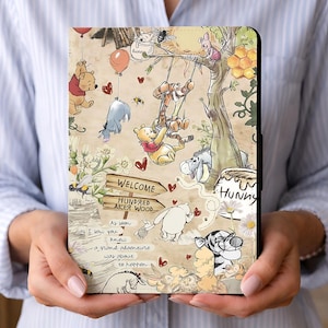 Può includere: Una custodia per tablet con un'illustrazione stravagante di Winnie the Pooh e amici. Il design include un albero, palloncini e il testo "WELCOME Hundred Aker Wood". La custodia è tenuta da una persona.