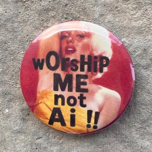 以下が含まれることがあります： 赤色の丸いバッジで、黒い文字で「wOrsHiP ME not Ai !!」と書かれています。バッジには、黄色のドレスを着た人のぼやけた画像があります。これはノベルティアイテムです。