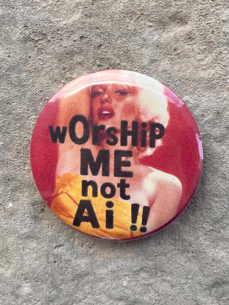 以下が含まれることがあります： 赤色の丸いバッジで、黒い文字で「wOrsHiP ME not Ai !!」と書かれています。バッジには、黄色のドレスを着た人のぼやけた画像があります。これはノベルティアイテムです。
