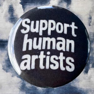 Pode incluir: Um bot&atilde;o redondo preto com as palavras "Support human artists" em branco. O bot&atilde;o tem um acabamento brilhante e est&aacute; sobre um fundo nublado azul e branco.