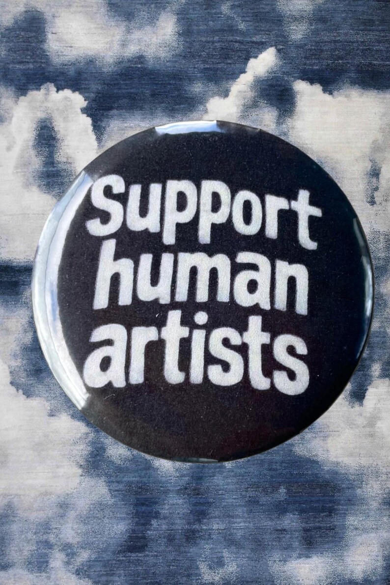 Pode incluir: Um bot&atilde;o redondo preto com as palavras "Support human artists" em branco. O bot&atilde;o tem um acabamento brilhante e est&aacute; sobre um fundo nublado azul e branco.
