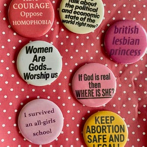 Puede incluir: Un conjunto de siete botones redondos con varios lemas y frases. Los botones son de color rosa, blanco y amarillo, con texto negro. Los lemas incluyen "Tener Coraje Oponerse a la Homofobia" y "¡Mantener el Aborto Seguro y Legal!"