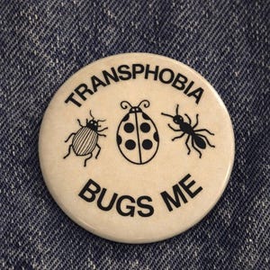 Puede incluir: Un botón redondo de color crema con las palabras "TRANSPHOBIA" y "BUGS ME" en negro, rodeando tres ilustraciones de insectos negros: un escarabajo, una mariquita y una hormiga. El botón está sobre un fondo de mezclilla azul.