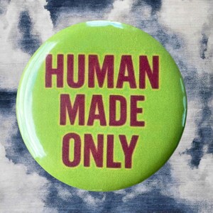 Pode incluir: Um bot&atilde;o redondo verde lim&atilde;o com o texto "HUMAN MADE ONLY" em letras bord&ocirc;. O bot&atilde;o est&aacute; sobre um fundo com um padr&atilde;o de nuvens azuis e brancas.