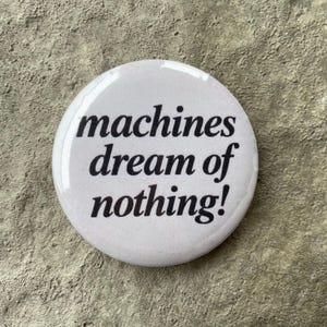 Może przedstawiać: Biała, okrągła przypinka z czarnym, pogrubionym napisem "machines dream of nothing!". Przypinka jest umieszczona na teksturowanym, szarym tle.
