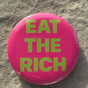 May include: A circular, pink badge with the green text "EAT THE RICH". The badge is set against a textured, grey background.