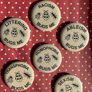 May include: A set of cream-coloured circular badges with black text and illustrations. Each badge displays the words "BUGS ME" alongside a ladybug, ant, and beetle. The badges address issues such as racism, littering, ableism, homophobia, transphobia, and sexism.