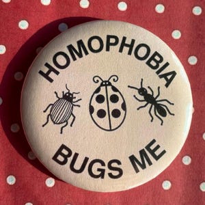 Puede incluir: Un botón redondo de color crema con las palabras negras "HOMOPHOBIA BUGS ME", que rodean tres ilustraciones de insectos negros: un escarabajo, una mariquita y una hormiga. El fondo es una tela roja con lunares blancos.