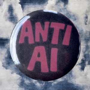 Pode incluir: Um bot&atilde;o redondo preto com as palavras "ANTI AI" em letras rosa e em negrito. O bot&atilde;o tem um acabamento brilhante e est&aacute; sobre um fundo de um padr&atilde;o nublado azul e branco.