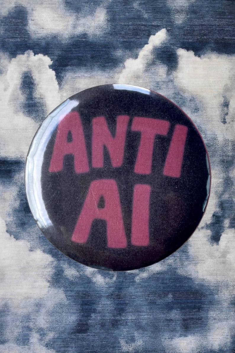 Pode incluir: Um bot&atilde;o redondo preto com as palavras "ANTI AI" em letras rosa e em negrito. O bot&atilde;o tem um acabamento brilhante e est&aacute; sobre um fundo de um padr&atilde;o nublado azul e branco.