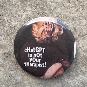 以下が含まれることがあります： 黒い背景に白い文字で「ChatGPT is not Your tHerapiSt!」と書かれた丸いボタン。金髪の人物のぼやけた画像が特徴です。ボタンはグレーのテクスチャのある表面にあります。