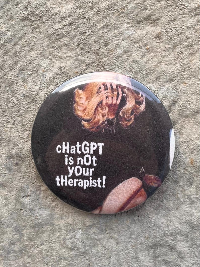 以下が含まれることがあります： 黒い背景に白い文字で「ChatGPT is not Your tHerapiSt!」と書かれた丸いボタン。金髪の人物のぼやけた画像が特徴です。ボタンはグレーのテクスチャのある表面にあります。