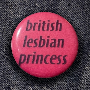 Può includere: Un bottone rotondo rosa con la scritta "british lesbian princess" in lettere viola scuro. Il bottone è posto su uno sfondo di denim blu.