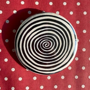 May include: A circular button featuring a black and white spiral design. The button is placed on a red background with white polka dots. The spiral pattern is centrally positioned, creating a visual vortex.