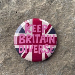 May include: A circular badge featuring the Union Jack flag in red, white, and blue. The words "KEEP BRITAIN DIVERSE" are printed in pink across the center. The badge is set against a textured, grey surface.