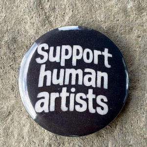 Op de afbeelding: Een ronde, zwarte knoop met de witte tekst "Support human artists". De knoop heeft een glanzende afwerking. De tekst is in een vetgedrukte, schreefloze lettertype. De knoop ligt op een getextureerd, grijs oppervlak.