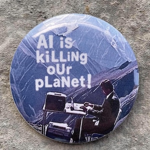 Pode incluir: Botão redondo com um design azul e branco. O texto "AI is killing our planet!" é exibido em branco, com uma silhueta de uma pessoa em uma mesa. O botão está em uma superfície cinza texturizada.