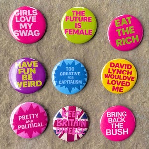 Puede incluir: Nueve botones redondos y coloridos con varios lemas. Los botones muestran frases como "Girls Love My Swag", "The Future is Female" y "Eat the Rich". Los botones están dispuestos sobre una superficie texturizada de tono neutro.