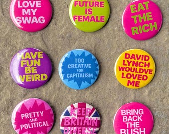 May include: Nine colorful pinback buttons with various slogans. The buttons are round and feature phrases like "Girls Love My Swag," "The Future is Female," and "Eat the Rich." The buttons are arranged on a textured, neutral-toned surface.