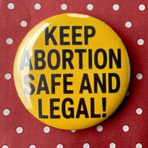 May include: A yellow button with the text "KEEP ABORTION SAFE AND LEGAL!" in black. The button is set against a red background with white polka dots.