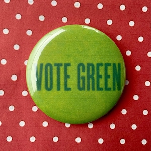 Op de afbeelding: Een ronde, groene button met de tekst "VOTE GREEN" in donkergroene letters. De button staat tegen een rode achtergrond met een wit stippenpatroon. De button heeft een glanzende afwerking.