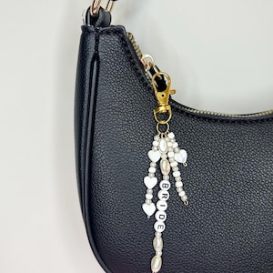 May include: A black handbag with a gold-colored zipper and hardware. A beaded charm with white heart and pearl accents hangs from the bag. The charm spells out the word "BRIDE" in white letter beads.