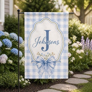 May include: A blue and white gingham garden flag with the name "Johnsons" in script. The flag features a large blue letter "J" and a decorative bow at the bottom. The flag is displayed on a black metal stand in a garden setting.