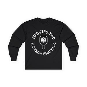 May include: Black long-sleeved shirt with a white graphic. The graphic shows a pickleball paddle inside a circle with the text "ZERO-ZERO-TWO" and "YOU KNOW WHAT TO DO".