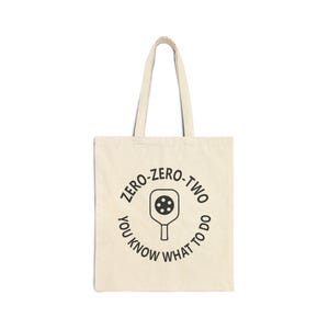 May include: Cream-colored tote bag with black text and a pickleball paddle graphic. The text reads "ZERO-ZERO-TWO YOU KNOW WHAT TO DO" in a circular design. The bag has two handles and is made of a canvas-like material.