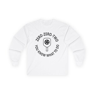 May include: White long-sleeved top with a circular graphic. The graphic features a pickleball paddle and the text "ZERO-ZERO-TWO YOU KNOW WHAT TO DO" in black. The shirt is made of a soft material.
