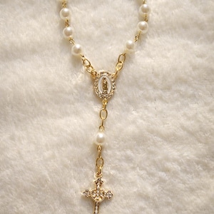 May include: A gold-colored rosary bracelet featuring faux pearls and a gold-tone cross pendant adorned with clear crystals. The bracelet includes a gold-tone centerpiece with a religious image.