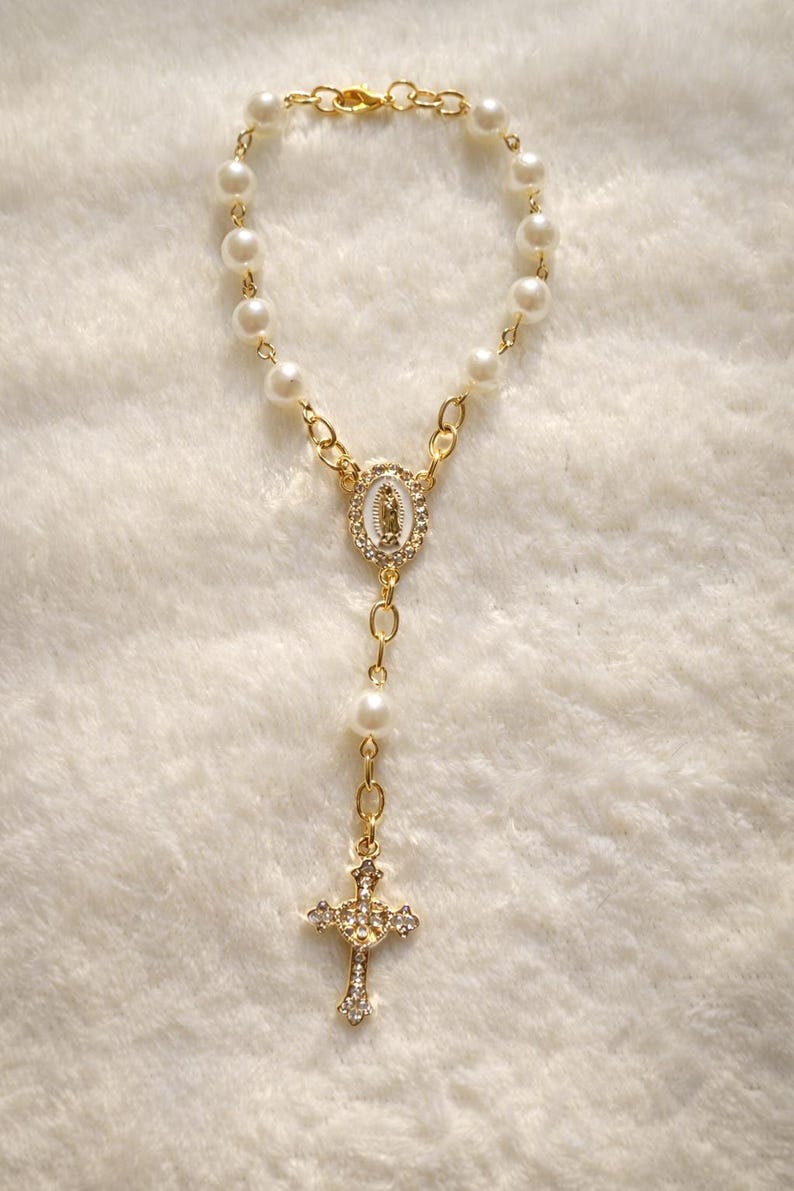 May include: A gold-colored rosary bracelet featuring faux pearls and a gold-tone cross pendant adorned with clear crystals. The bracelet includes a gold-tone centerpiece with a religious image.