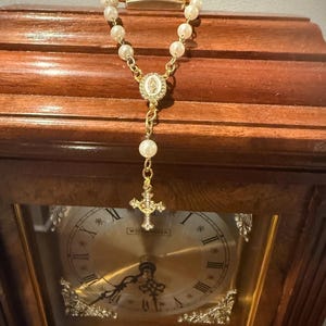 May include: A gold-toned rosary with faux pearl beads hangs in front of a wooden clock. The rosary features a cross pendant and a central medallion. The clock has a gold-colored face with Roman numerals and decorative accents.