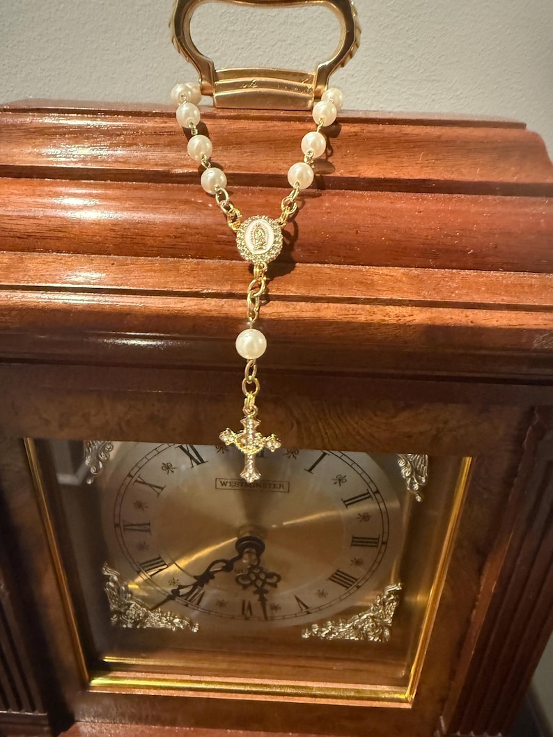 May include: A gold-toned rosary with faux pearl beads hangs in front of a wooden clock. The rosary features a cross pendant and a central medallion. The clock has a gold-colored face with Roman numerals and decorative accents.