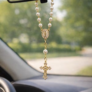 May include: A car rosary with faux pearl beads and gold-tone accents. The rosary features a cross pendant with clear crystal-like embellishments and a central medallion with a religious figure.